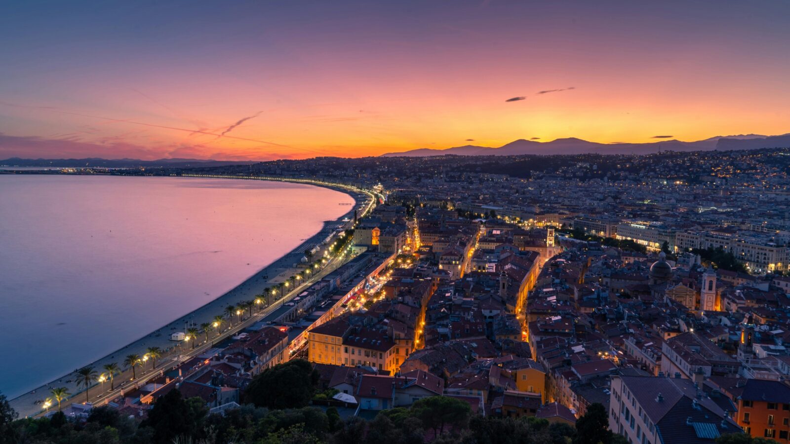 Uitzicht over Nice bij zonsondergang, met de kustlijn, verlichte gebouwen en een paars-oranje lucht boven de stad en de zee.