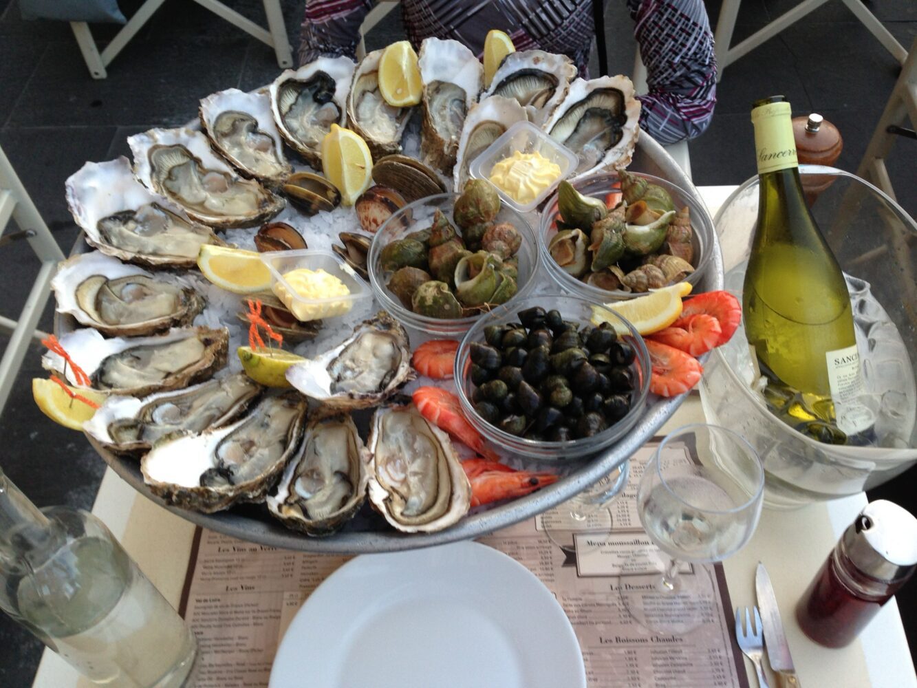 Een schaal met zeevruchten, waaronder oesters, garnalen en slakken, geserveerd met citroen en een fles witte wijn.